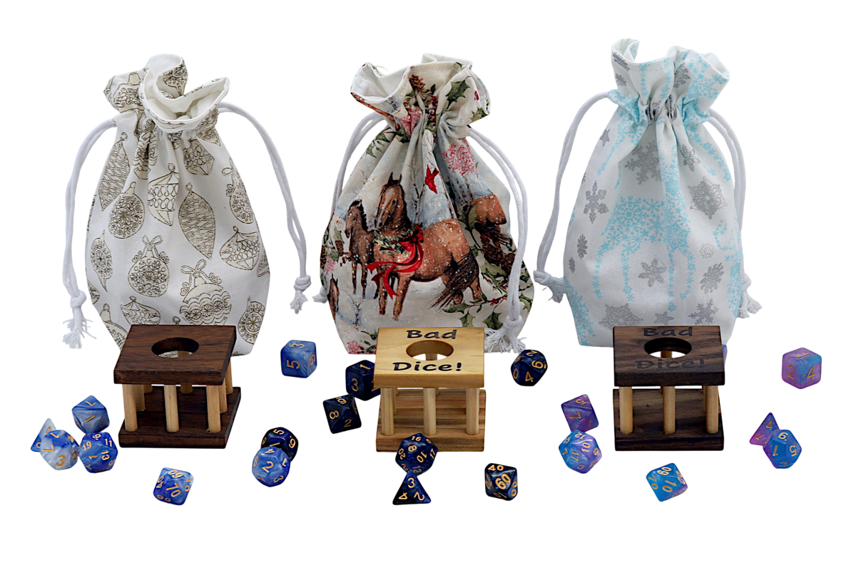 Three fabric drawstring dice pouches with white cords are arranged behind small wooden dice cages, with assorted polyhedral gaming dice scattered in front on a white background. The left pouch is white with gray ornament motifs, the center pouch is cream with a red and brown horse-and-bow print, and the right pouch is white with light blue snowflake and deer motifs.